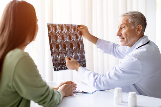 Senior male doctor in white coat showing Xray image to female patient during consultation in bright clinic office, medical diagnosis concept