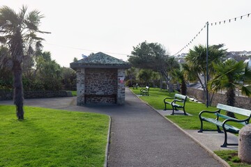A paved path runs through a park lined with palm trees and green benches. A stone bus shelter sits on the left in Perranporth - Cornwall - UK