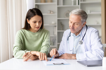 Pregnant woman and senior male doctor reviewing medical records together during prenatal consultation in bright clinic office, pregnancy healthcare care