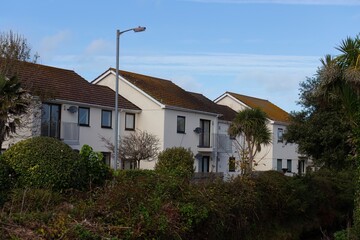 A row of white houses with brown roofs are shown from a distance. Green shrubbery sits in front of the houses in Perranporth - Cornwall - UK