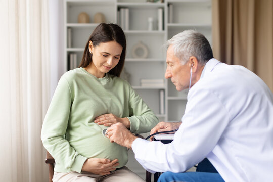 Senior male doctor using stethoscope to examine pregnant womans belly during prenatal checkup in clinic, maternity healthcare and pregnancy care concept - Powered by Adobe