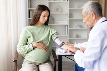 Senior male doctor measuring blood pressure of pregnant woman during prenatal examination in clinic, healthcare monitoring and maternity checkup concept