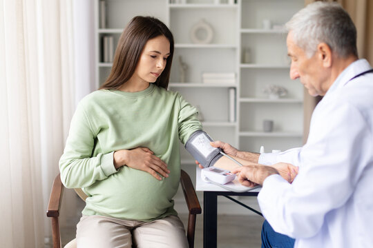 Senior male doctor measuring blood pressure of pregnant woman during prenatal examination in clinic, healthcare monitoring and maternity checkup concept