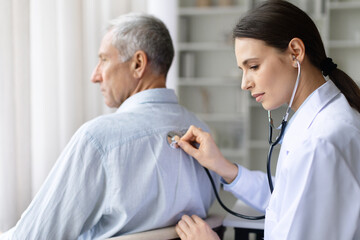 Fototapeta premium Female doctor in white coat examining senior male patients back with stethoscope during medical checkup in bright clinic office, professional healthcare service