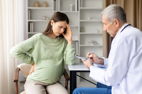 Pregnant woman suffering headache and back pain consulting senior male doctor in clinic office, pregnancy discomfort and prenatal healthcare concept