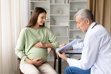 Smiling pregnant woman talking with senior male doctor in clinic during prenatal consultation, maternity healthcare and pregnancy monitoring concept