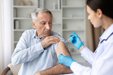 Female doctor in white coat giving vaccine injection to senior male patient in clinic, immunization and healthcare prevention concept