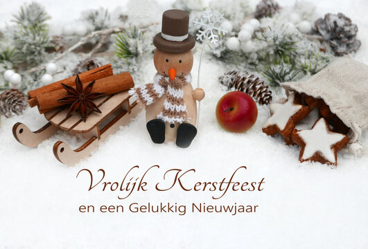 Kerstkaart: Kerstversiering met kaneelsterren, appels en een houten sneeuwpop. De Nederlandse tekst "Vrolijk Kerstfeest en een Gelukkig Nieuwjaar" staat in de sneeuw.