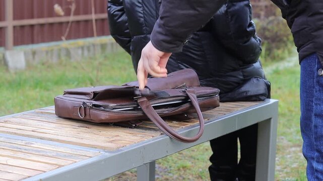 In a public area, an individual approaches a bag placed on a bench while another person sits unaware. The setting is outdoors, suggesting a possible theft of a laptop.