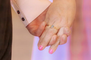 Intimate close-up of a couple holding hands, highlighting a gold engagement ring with a gemstone on a woman's hand. Pink nail polish and soft lighting suggest a romantic moment.