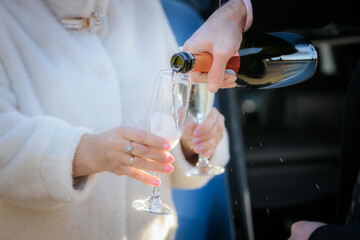 person pouring champagne into a flute held by a woman in a white coat, wearing an engagement ring. Scene suggests a romantic or festive occasion.