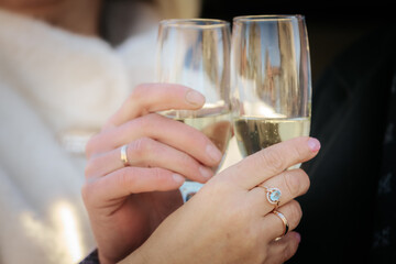 A romantic close-up of a couple clinking champagne glasses, both wearing wedding rings. The focus is on their hands and sparkling wine, suggesting celebration or engagement.