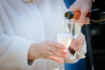 person pouring champagne into a flute held by a woman in a white coat, wearing an engagement ring. Scene suggests a romantic or festive occasion.