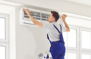 Male repairman checks filter and inspects air conditioning unit in residential building. Man in overalls stands on ladder and removes cover of air conditioner. Air conditioner maintenance concept. © Studio Romantic