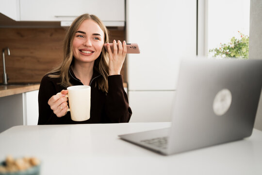 Young woman business worker drinking tea, using laptop listening to voice message on smartphone working at home office