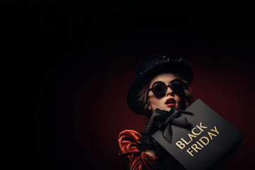 femme robe rouge et chapeau melon noir, lunettes de soleil, et gants noirs avec un sac de course noir texte doré en anglais "Black Friday" fond rouge espace négatif copyspace soldes fin novembre