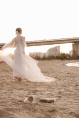 Naklejka premium Bride Dancing on a Sandy Beach at Sunset with Flowing Veil and Urban Bridge in the Background