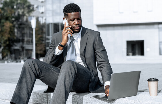 A man in a suit sits on a concrete step in an urban area. He is using a laptop while speaking on his phone. The scene captures a moment of multitasking in a modern environment. - Powered by Adobe