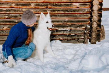 A woman in a knit hat and winter clothes sits beside a white dog in the snow, in front of a rustic wooden cabin. Peaceful outdoor winter moment with a loyal pet.