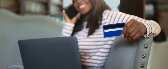 A woman sits in a comfortable chair, smiling and talking on her phone. She holds a credit card in one hand while typing on her laptop, engaged in online shopping in her cozy living room.