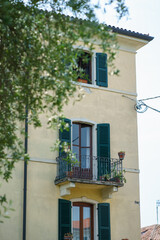 A traditional Italian home with green shutters, a cozy balcony full of plants, and soft daylight illuminating its warm yellow facade in Bardolino.