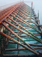 Fototapeta premium Weathered, rusty support structure of a pier or jetty extending into foggy turquoise water. The metal beams are heavily covered in rust, algae, and barnacles. The fog creates a moody background