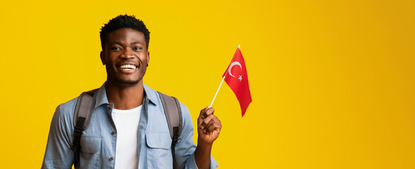 A cheerful young man holds a small Turkish flag, smiling widely. He wears a casual outfit and a...