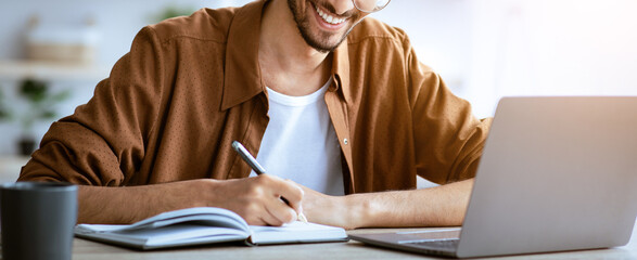A young man smiles while writing notes in a notebook during a study session. He sits at a desk with a laptop and a coffee cup, creating a cozy and productive atmosphere.