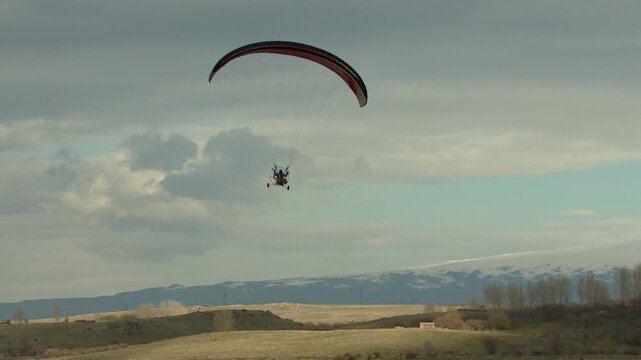 A motorized paraglider (paramotor/powered parachute) flies across a cloudy sky, towing a white advertising banner. Recreation, extreme sport, and aerial advertising activity.