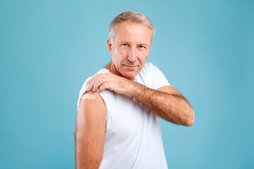 A happy senior man smiles while revealing his shoulder with a bandage after receiving the Covid-19 vaccine. He poses confidently against a blue background, celebrating his vaccination.