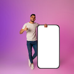 Happy millennial Arab man stands beside a giant smartphone with a blank screen, giving a thumbs up...