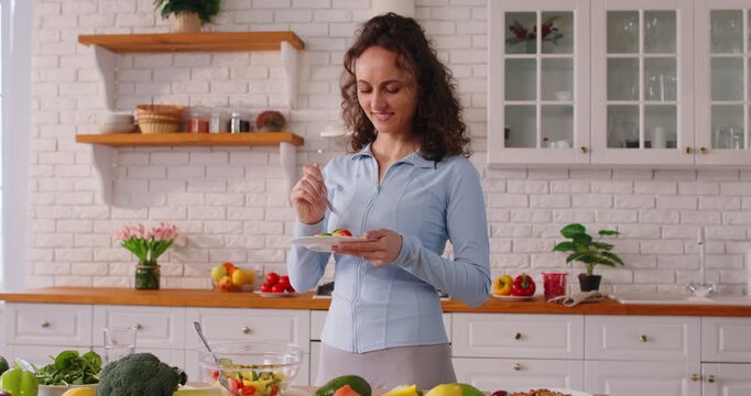 Slim woman tasting delicious vegetable salad she prepared, posing at kitchen, keeping to healthy nutrition, having lunch, enjoying fresh veggies. People, nutrition and dieting concept