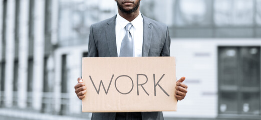 A man dressed in a professional suit stands outdoors in a city environment, holding a large sign that reads work. The scene conveys a sense of job searching and ambition during daylight.
