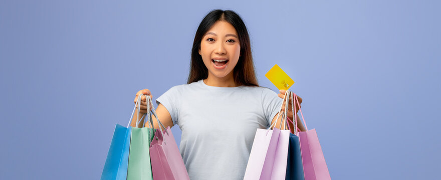 A young woman smiles joyfully while holding multiple colorful shopping bags. She shows a credit card, celebrating her shopping experience against a light blue background. - Powered by Adobe