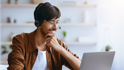 A young man is smiling while working on his laptop at home. He wears a headset and looks engaged in an online meeting, surrounded by a bright workspace and plants.