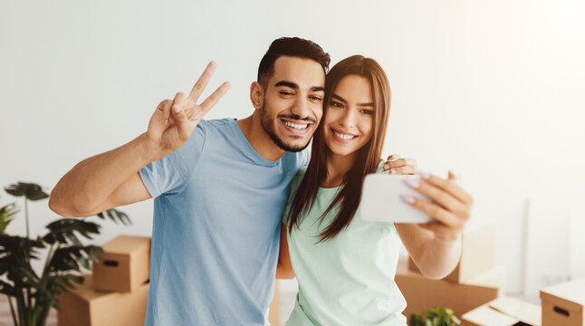 A young couple smiles and poses for a selfie in their new apartment. They are surrounded by cardboard boxes and indoor plants, celebrating their recent move together.
