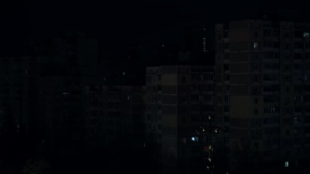 A dramatic timelapse capturing a city block in Ukraine during a blackout with flickering lights from windows illuminating the darkness, showcasing the silhouette of residential buildings