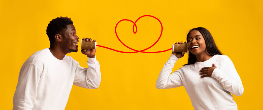 Two happy individuals enthusiastically talk to each other using tin can telephones. The bright yellow backdrop enhances their joyful expressions, creating a fun atmosphere filled with love.
