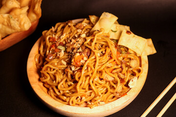 Spicy fried noodles with wonton crackers in a wooden bowl, isolated on a black background with a wooden plate and traditional chopsticks