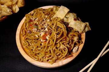 Spicy fried noodles with wonton crackers in a wooden bowl, isolated on a black background with a wooden plate and traditional chopsticks
