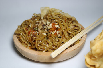 A bowl of spicy fried noodles with wonton crackers in a bowl, isolated on a white background with a wooden plate and traditional chopsticks