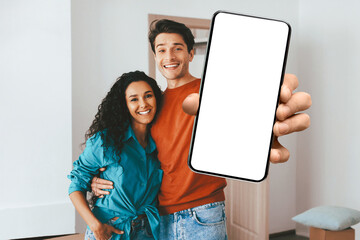 A couple stands close together in a well-lit room, smiling at the camera. One partner holds a smartphone to showcase its screen while they both appear happy and relaxed.
