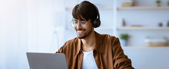 A young man with headphones sits in a bright home office. He focuses on his laptop with a smile, showing engagement in his work. The atmosphere is relaxed and productive.