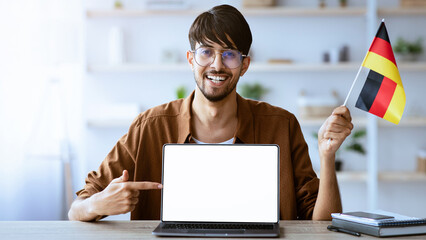 A young man is sitting at a desk in a well-lit room, joyfully pointing at a blank laptop screen while holding a small German flag. The background features plants and books.