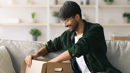 A young man sits on a couch and smiles as he opens a box. Natural light fills the room, creating a warm atmosphere. He anticipates discovering what is inside the package.