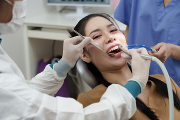 Fototapeta premium Asian adult female patient smiling while receiving oral cleaning treatment in dental clinic, dentist uses scaler and suction equipment, dental assistant present in background during procedure