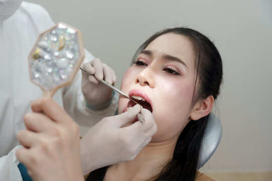 Asian woman holding mirror sitting in dental chair while dentist uses scaler and explorer to examine teeth during cleaning treatment at dental clinic with proper protective gear