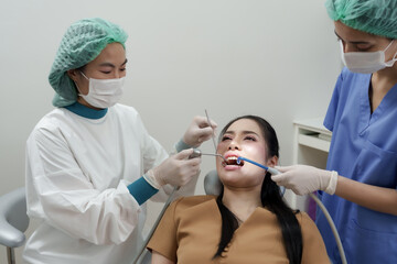 Asian adult woman being treated on dental chair as dentist uses dental mirror and assistant holds curing light, preparing composite material for tooth restoration in modern clinical setting