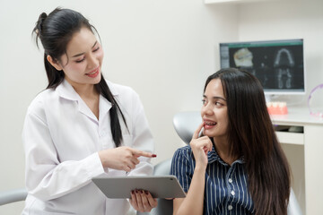 Asian female dentist pointing at dental model while explaining oral hygiene tips to multiracial female patient with braces in dental chair inside modern clinic with dental X-ray displayed behind