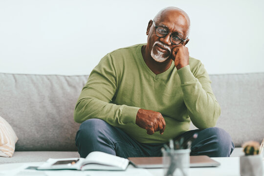An elderly man sits comfortably on a couch, looking thoughtful. He is dressed in a green sweater and is in a relaxed living room setting, surrounded by books and plants.